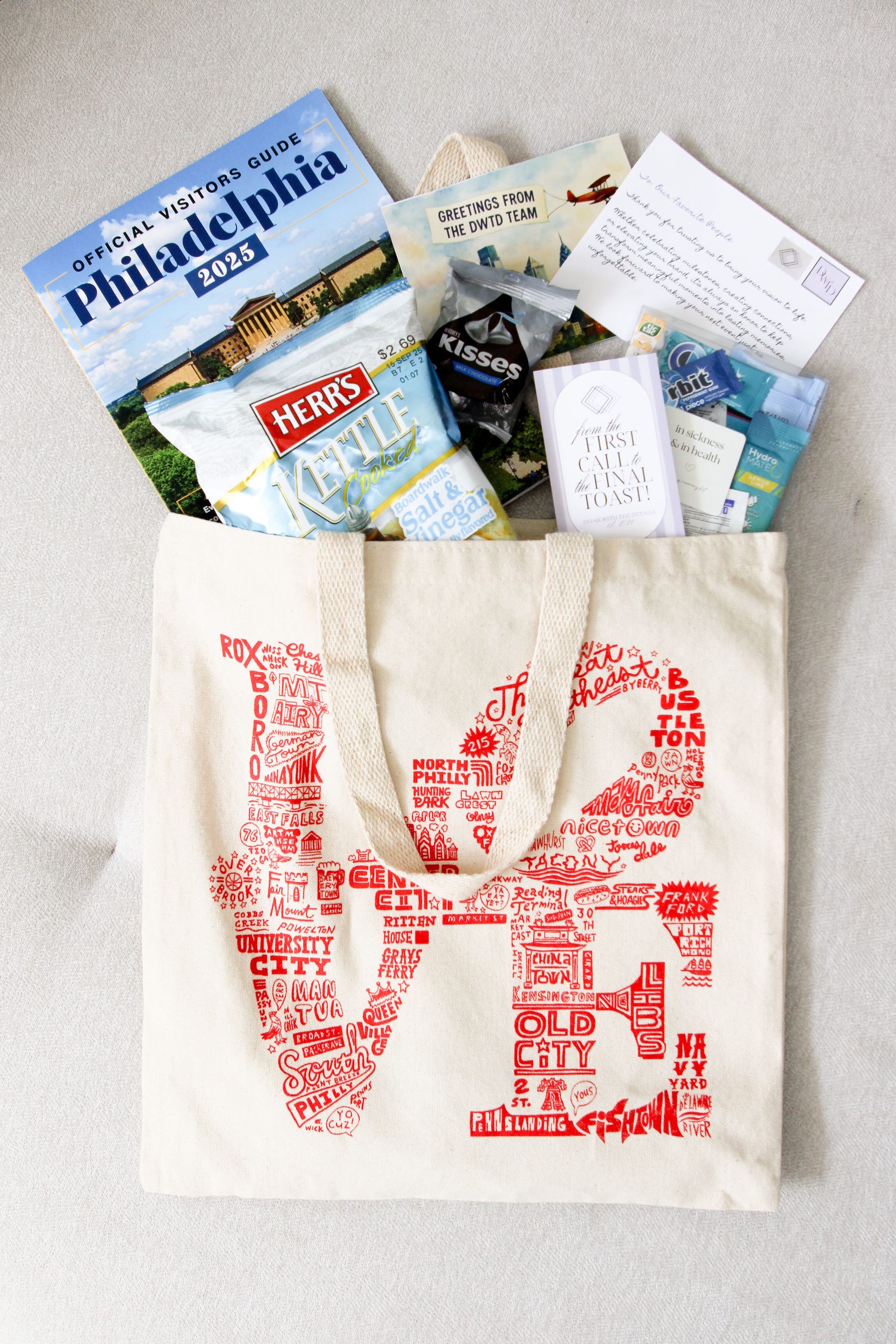 Canvas tote bag printed with a red Philadelphia LOVE design made of neighborhood names, filled with an official Philadelphia visitors guide, Herr’s kettle chips, Hershey’s Kisses, mints, gum, hydration packets, and a “Greetings from the DWTD Team” postcard with a welcome note.